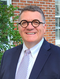 Man smiling and wearing blue suit jacket with a white shirt and pink and blue striped tie, and black round eye glasses.
