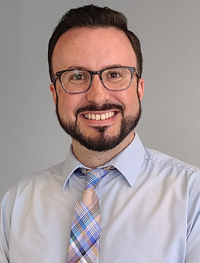 Professional young man with dark hair, beard, mustache, and eye glasses wearing a button-down shirt and multi-color plaid tie.