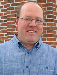 Smiling man wearing metal rimmed eye glasses and a blue button-down collared shirt standing in front of a brick wall.